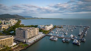A marina with multiple boats docked, surrounded by modern buildings and a clear view of the sea and distant mountains. - Belle Escapes - 405 Harbour Lights (Cairns City)