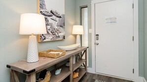 Un pasillo con una consola de madera, dos lámparas de pared, una fotografía enmarcada de una playa y una puerta blanca con ranura para la tarjeta de seguridad.