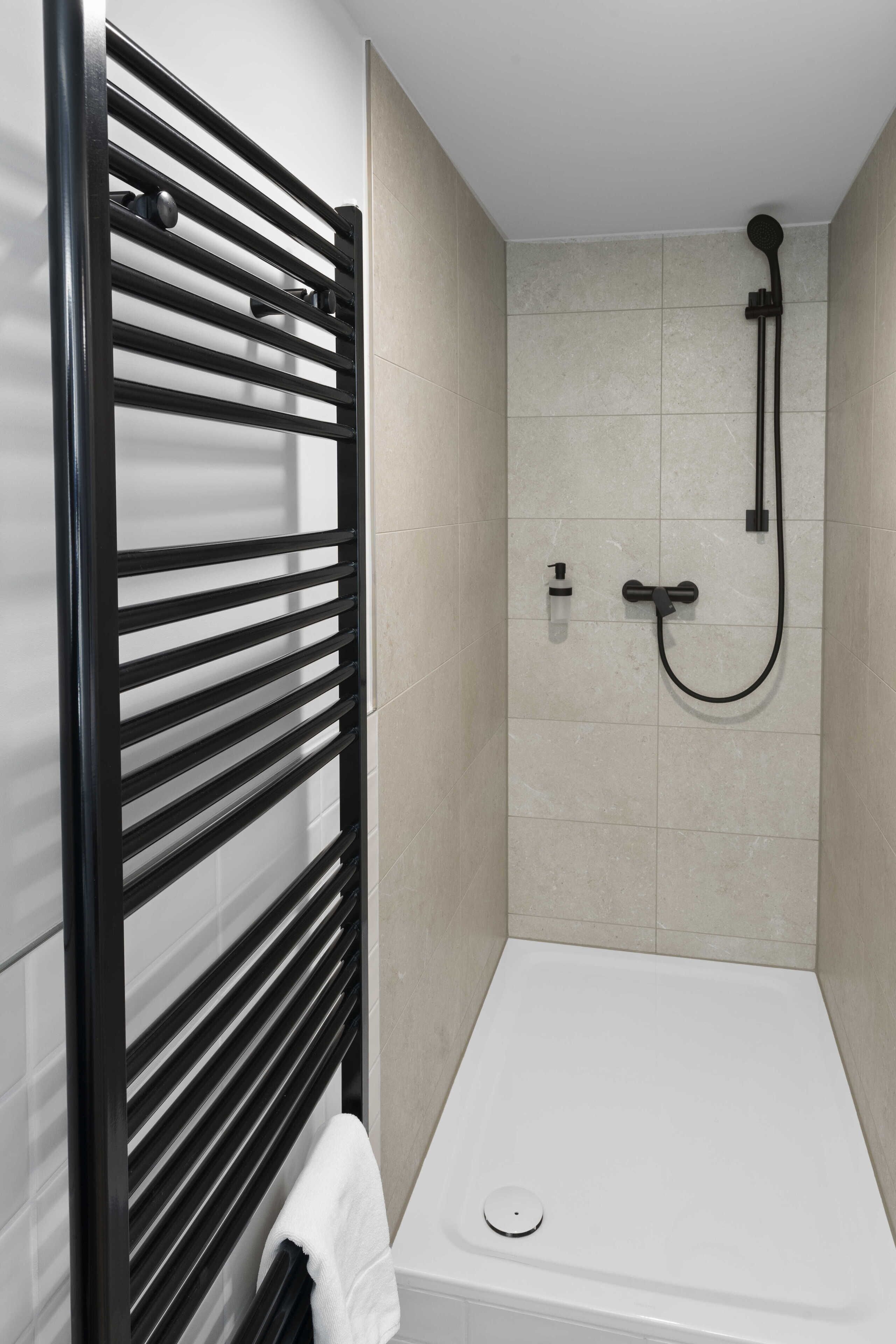 Une salle de bain moderne avec un porte-serviettes noir, une douche équipée d’un pommeau de douche à main et une baignoire blanche.