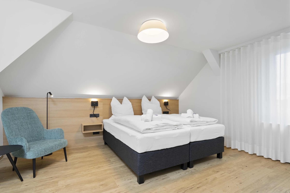 A modern hotel room with a bed, a chair, a bedside table, and a ceiling light.
