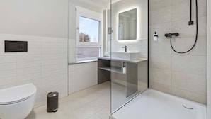 A modern bathroom with a toilet, a glass-enclosed shower, a sink, and a window. - Hotel Garni Traube (Friedrichshafen)