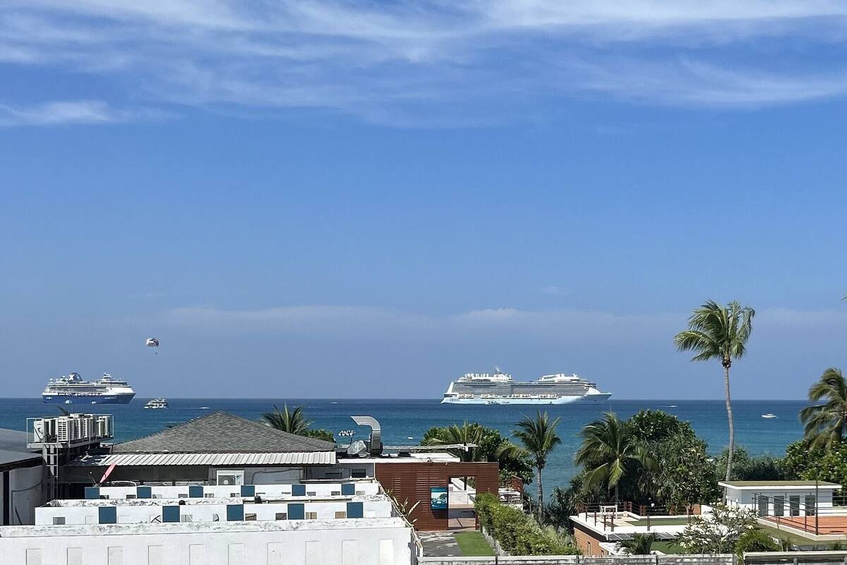 A coastal view with a cruise ship in the distance, palm trees, and buildings with a view of the ocean.