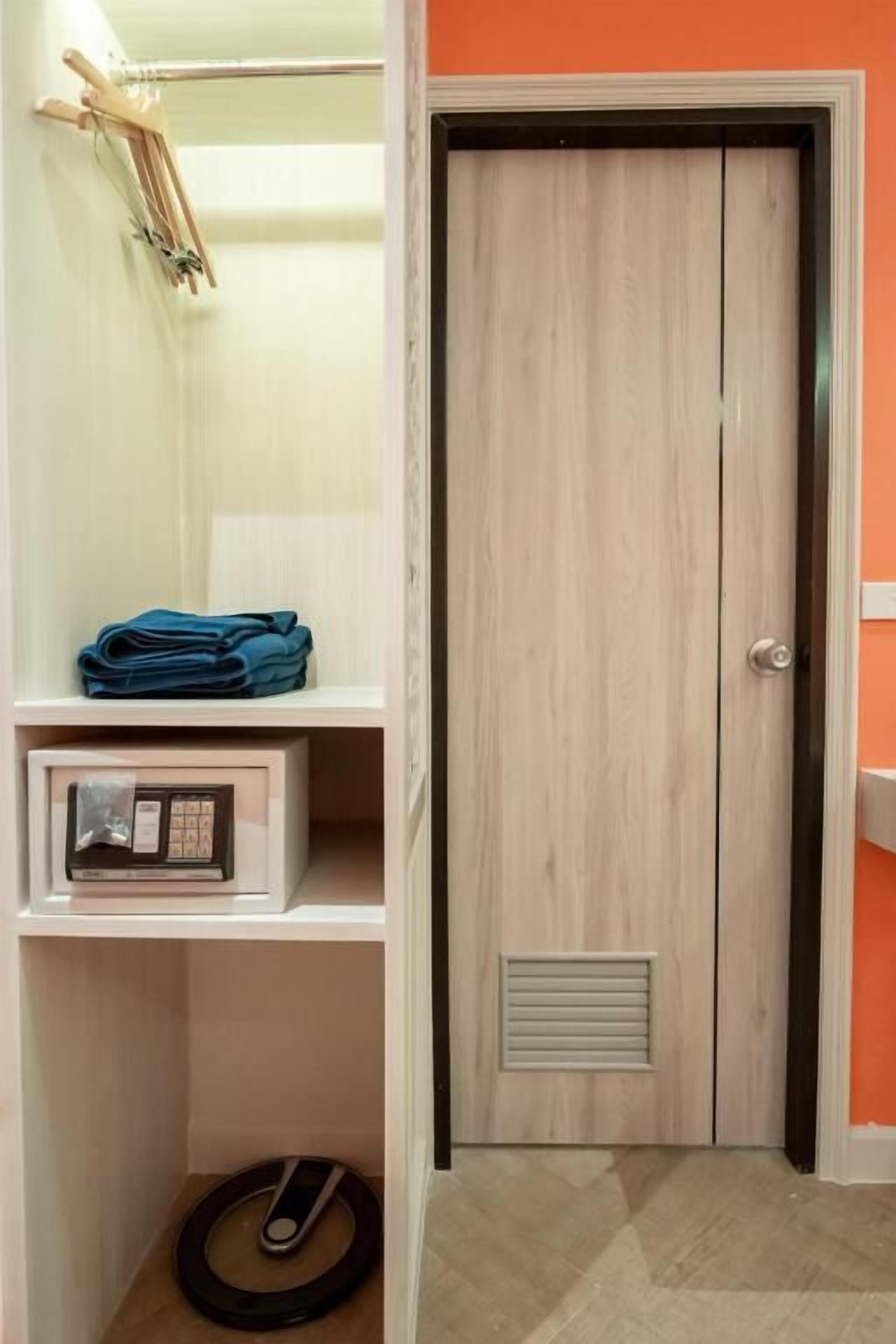 Une pièce avec une porte en bois, une petite étagère où sont pliés des vêtements, une radio et un objet circulaire sur le sol.