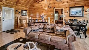A log cabin interior with a wooden ceiling, a sofa with animal-themed cushions, a dining table with chairs, and a fireplace.