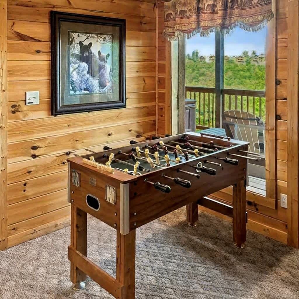 Ein Zimmer mit Holzboden, einer Tischfußballplatte, einem gerahmten Bild mit Wildtieren und Blick auf Grün durch ein Fenster.