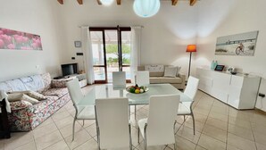 A modern living room with a glass dining table, white sofas, and a kitchen area with a white cabinet. - Villa Fiori with pool by Wonderful Italy (Olbia)