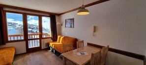 A room with a wooden floor, a sofa, a table with chairs, a hanging lamp, and a view of snow-covered mountains through large windows.