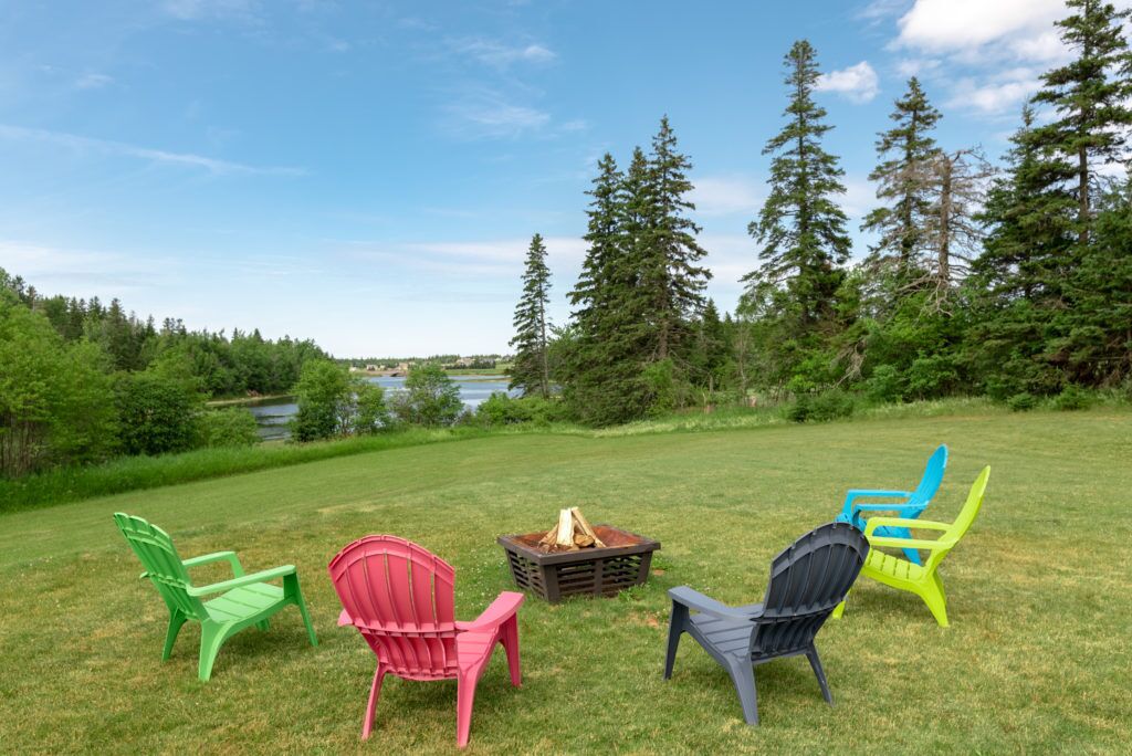 A grassy outdoor area with four Adirondack chairs arranged in a semi-circle, a fire pit in the center, and a lake with trees in the background.