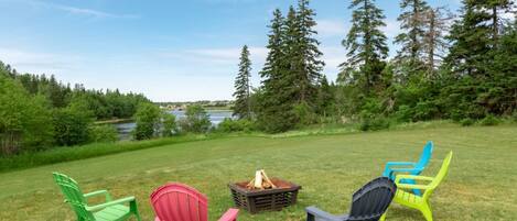 A grassy outdoor area with four Adirondack chairs arranged in a semi-circle, a fire pit in the center, and a lake with trees in the background.