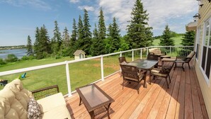 A wooden deck with outdoor furniture, a white railing, and a view of a lake and forest. - Storybook Island Cottage at The Gables of PEI (Stanley Bridge)