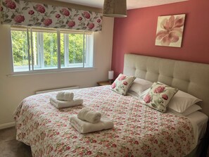 A bedroom with a large bed, floral bedding, a window with curtains, and a wall-mounted lamp. - Ludlow House (Ludlow)