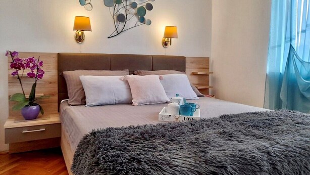 Une chambre à coucher moderne avec un grand lit, deux lampes de chevet, un élément décoratif mural, une table de chevet avec un vase de fleurs et un tapis moelleux.