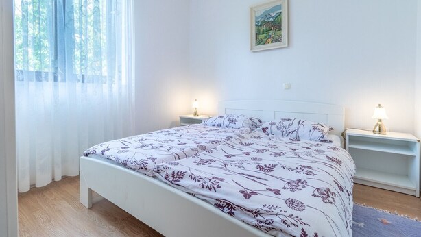 Une chambre à coucher avec un lit, deux tables de chevet, une commode, une fenêtre avec des rideaux légers et un tableau au mur.