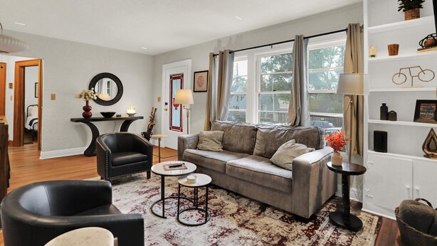 A living room with a grey sofa, black armchairs, and a coffee table. There is a bookshelf with decorative items and a window with curtains. - Hoosier Haven Spacious Retreat (Bloomington)
