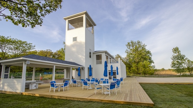 A white building with a tower, a covered patio with blue chairs and umbrellas, and a grassy deck.