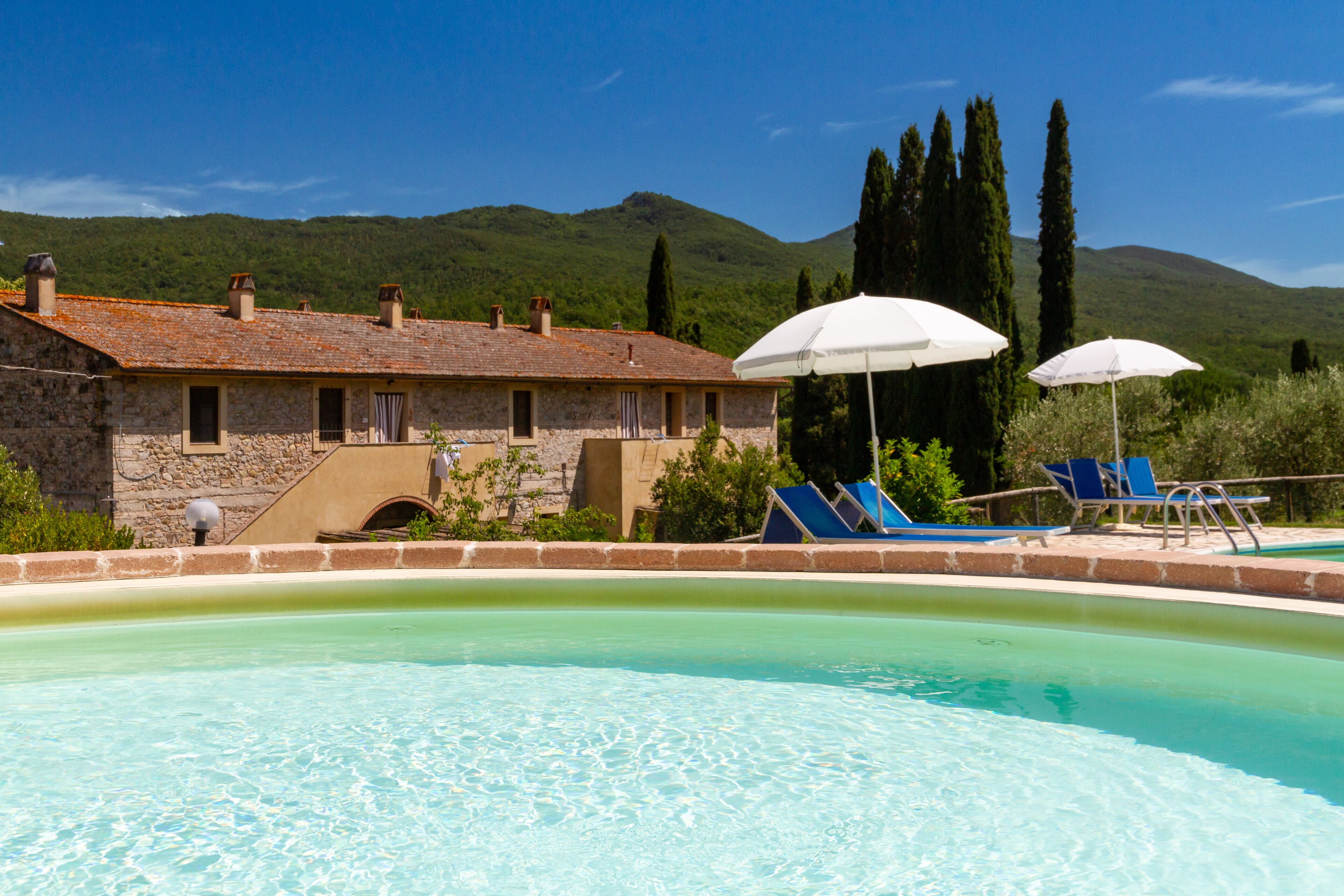 A pool area with a stone building, two umbrellas, and blue lounge chairs.