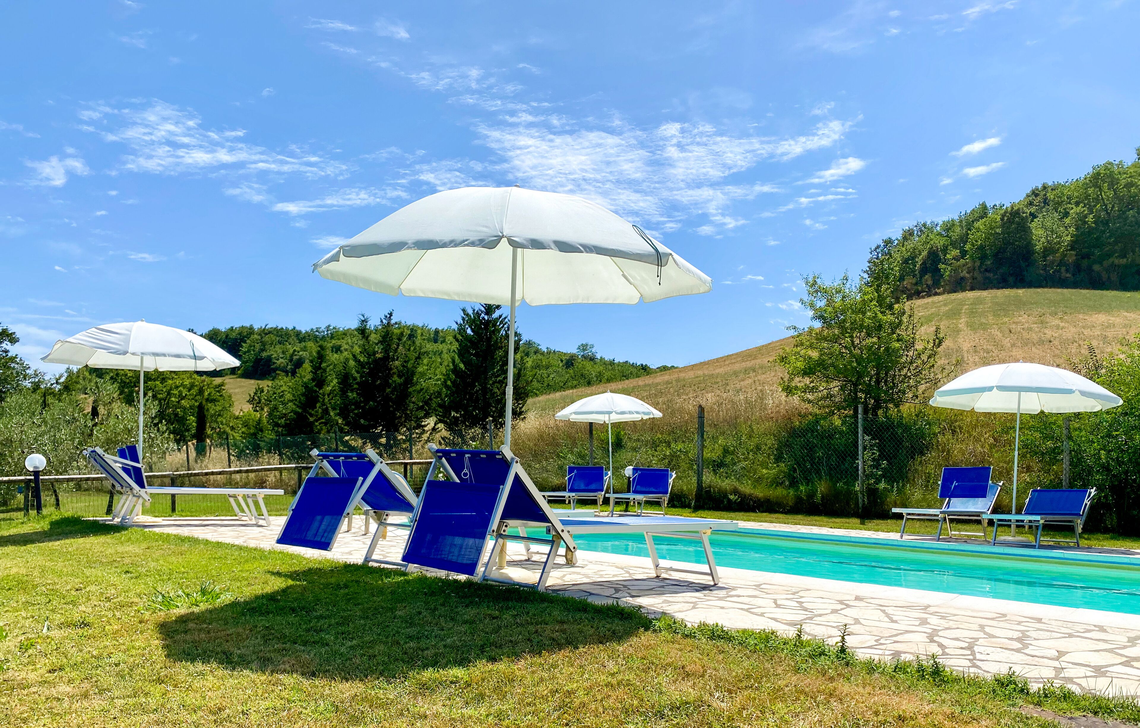 Uma área de piscina com espreguiçadeiras azuis e guarda-sóis brancos, cercada por gramado e árvores.