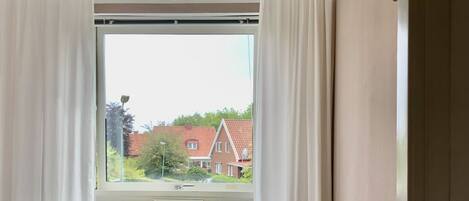 Une chambre à coucher avec un lit, un radiateur, une fenêtre avec des rideaux et une vue sur les maisons à l’extérieur.
