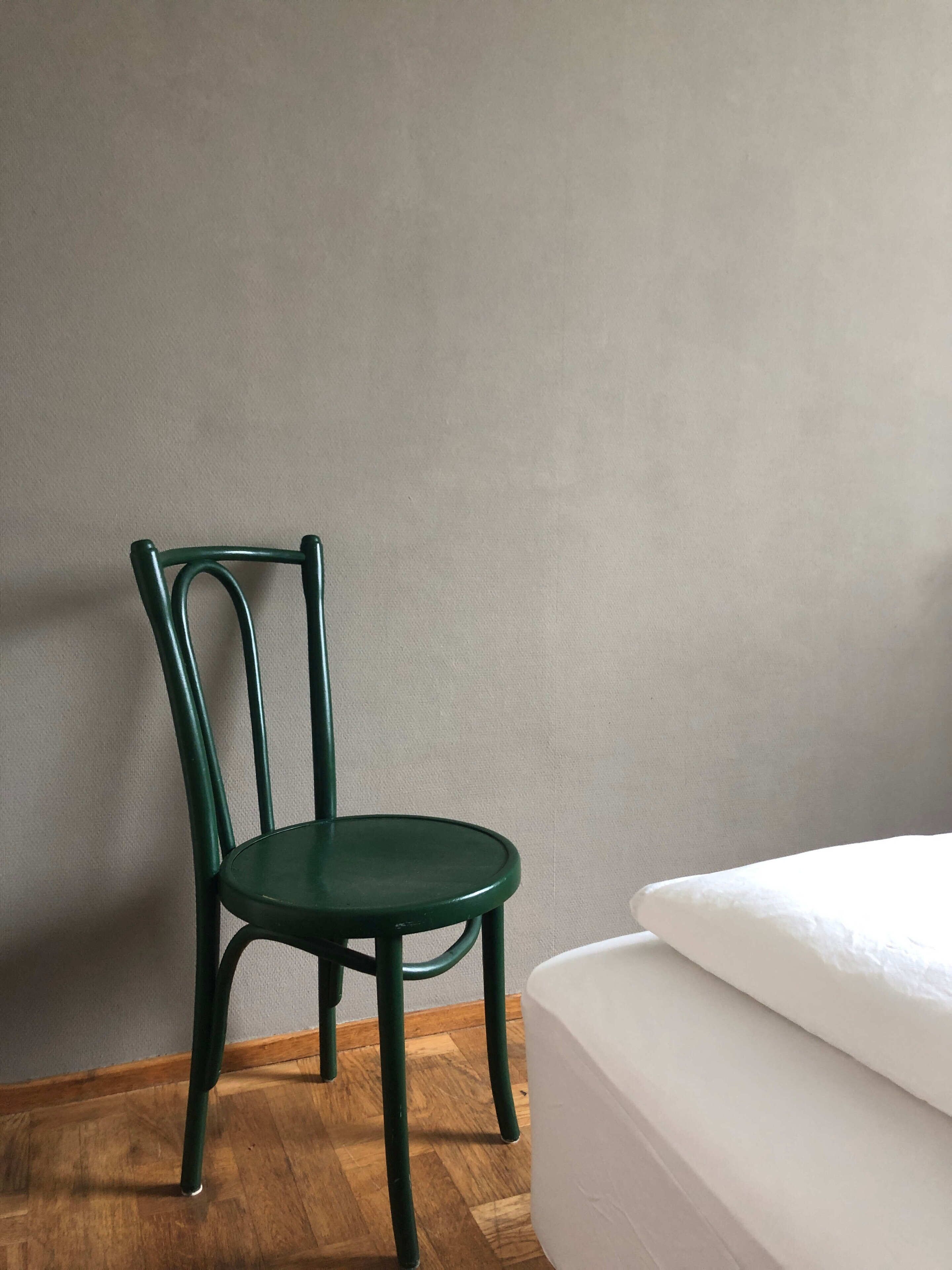 A room with a green metal chair, a wooden floor, and a white bed against a grey wall.
