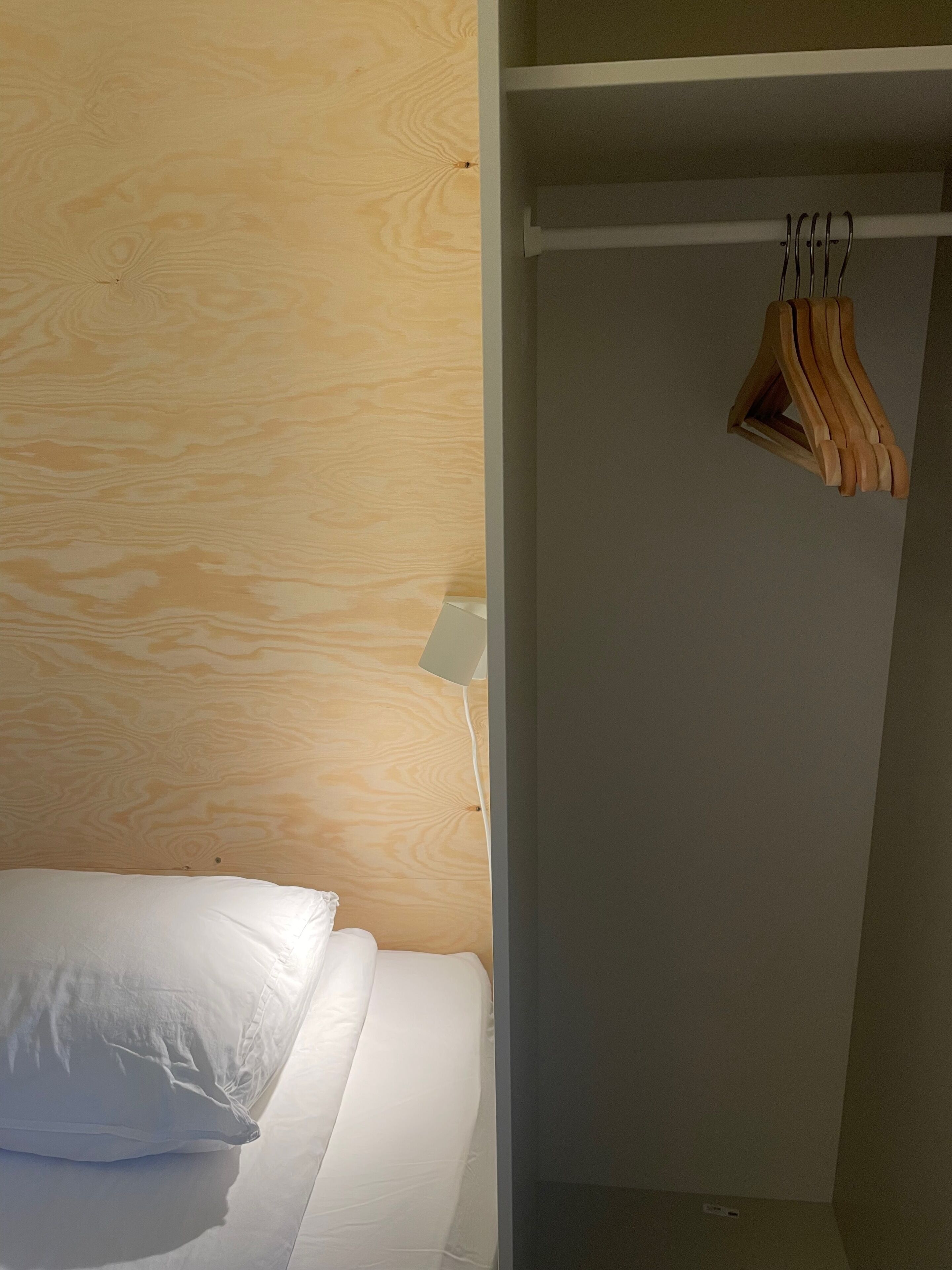 Ett sovrum med en sänggavel i trä, en vit säng med en kudde och en upphängd trähylla.