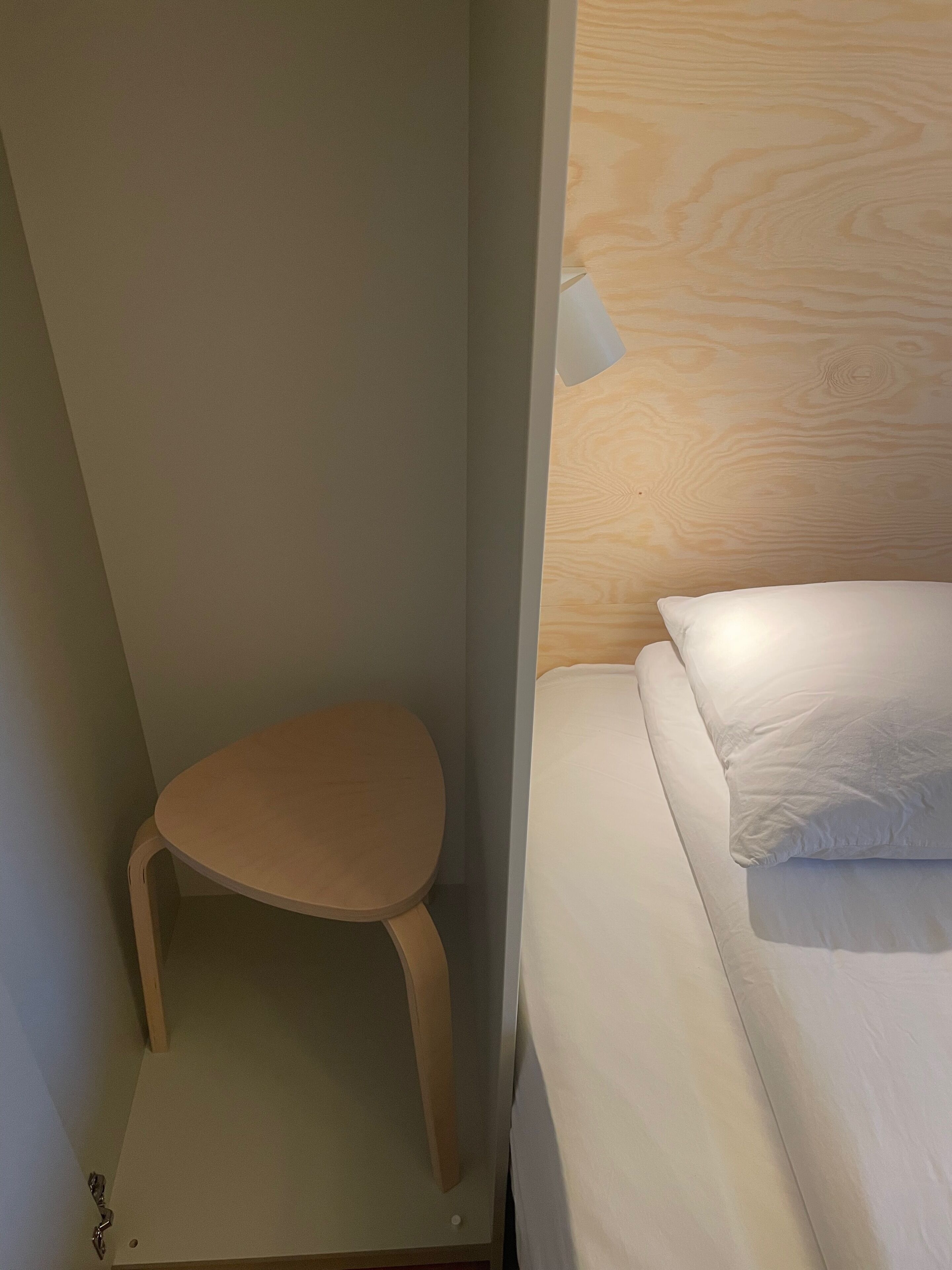 Una habitación pequeña y cerrada con una cama individual, un taburete de madera y un panel decorativo de madera en la pared.