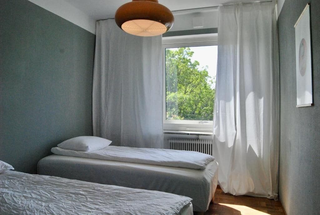 Un dormitorio con dos camas, una ventana con cortinas, un radiador y una lámpara colgante.