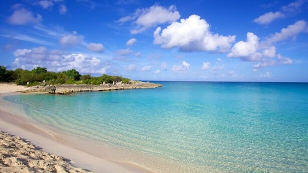 A beach with clear turquoise water, a sandy shore, and a rocky outcrop with vegetation. - Blue Maho Sxm (Sint Maarten)