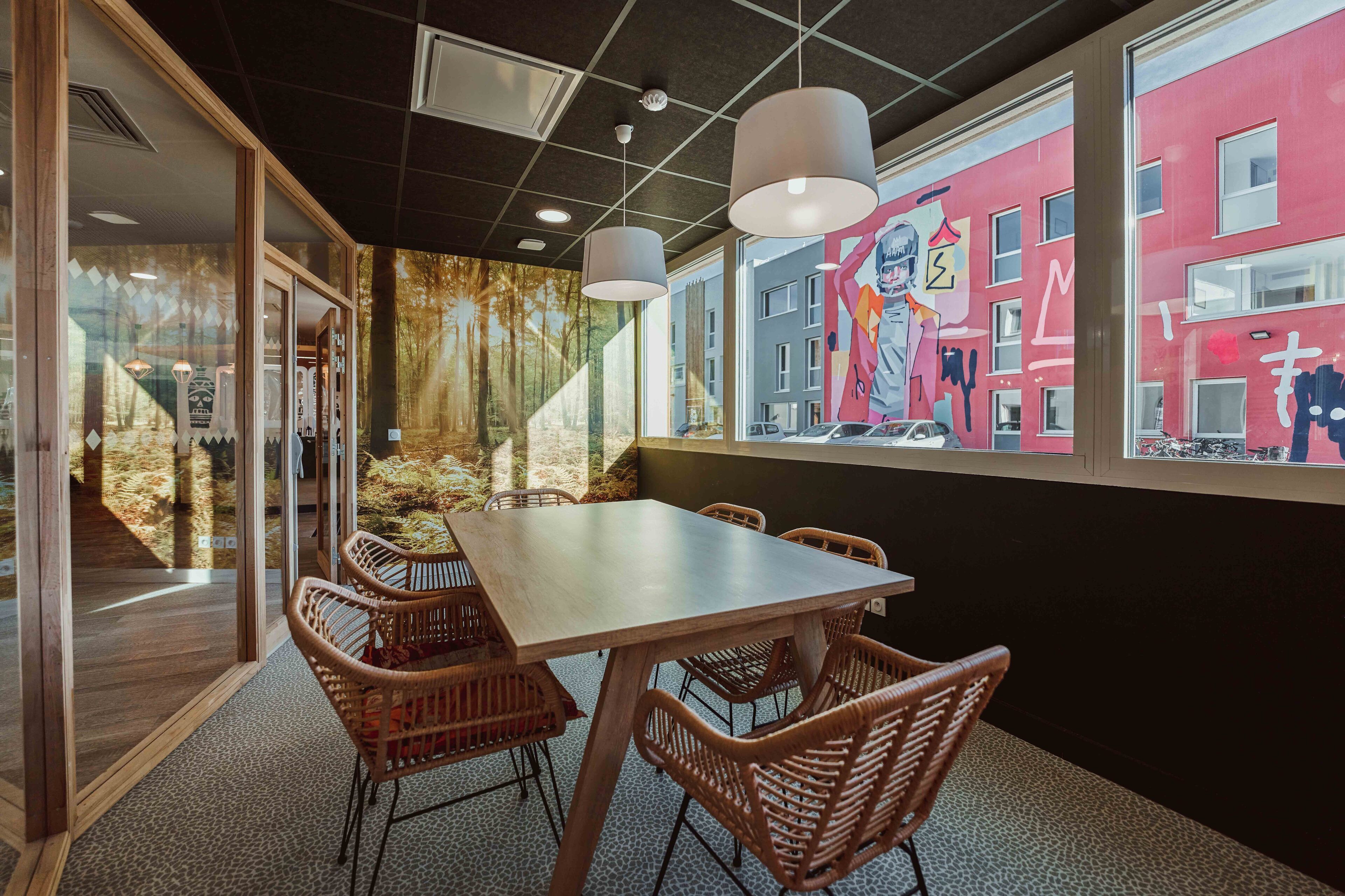 Een congreszaal met een grote tafel en rieten stoelen, een muurschildering aan de wand, en uitzicht op een rood gebouw.