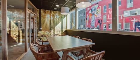 A conference room with a large table and wicker chairs, a mural on the wall, and a view of a red building outside.