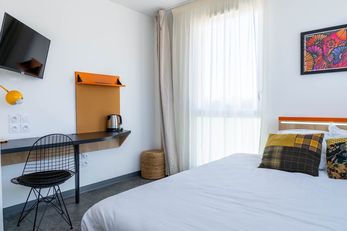 A hotel room with a bed, a desk with a chair, a television, and a window with curtains.