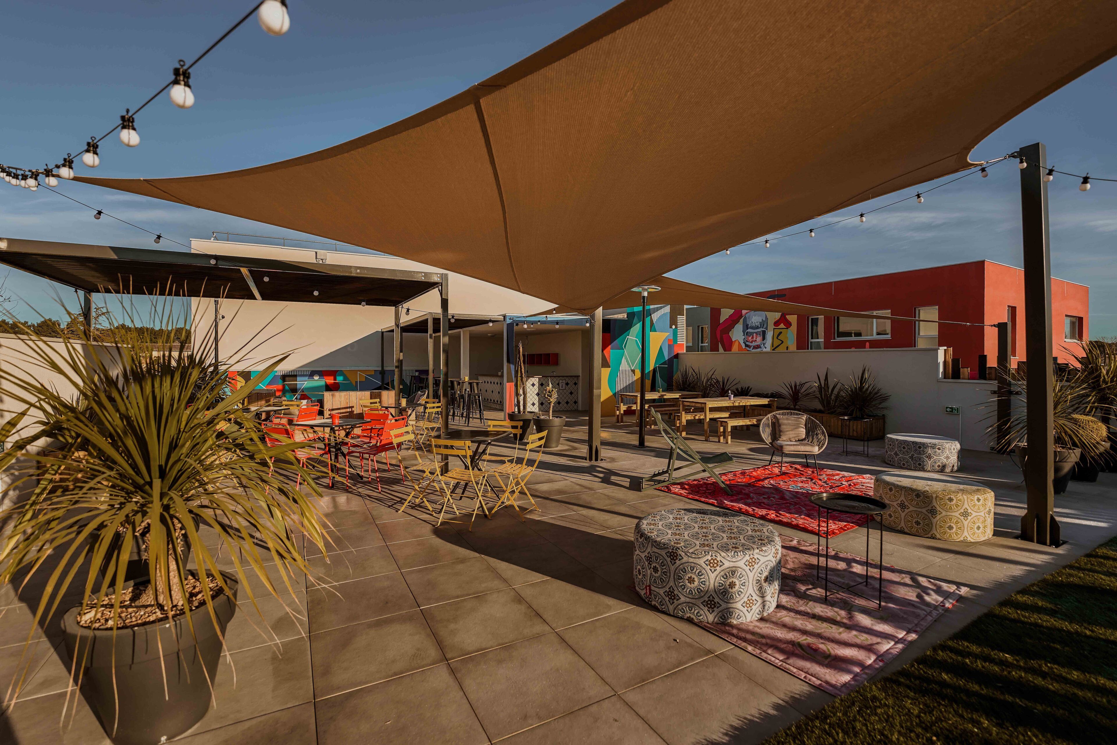 An outdoor patio area with seating arrangements, a large shade structure, and a building with colorful artwork in the background.