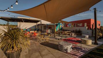 Un patio al aire libre con arreglos de asientos, una gran estructura de sombra y un edificio con arte colorido al fondo.