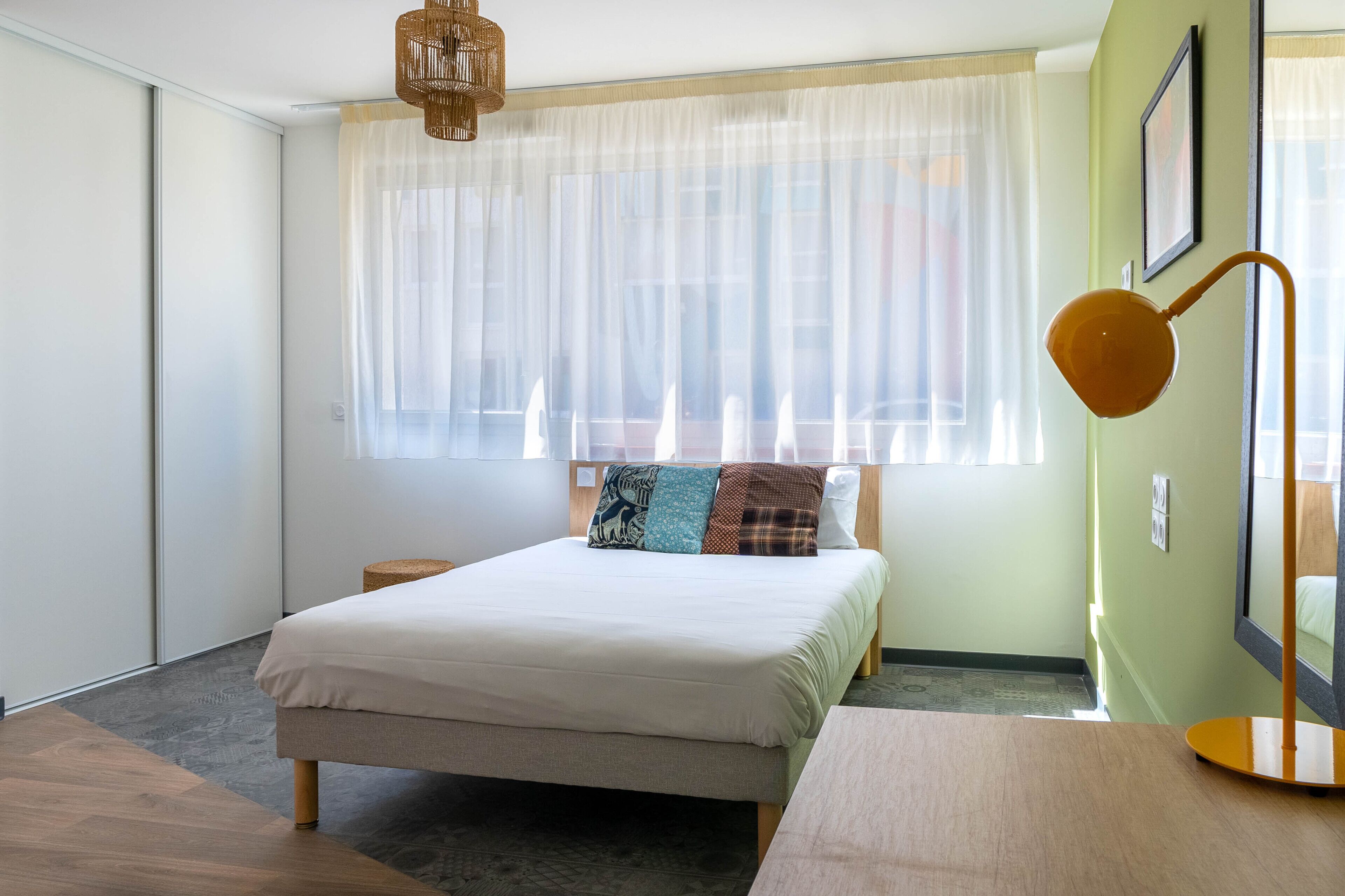 A bedroom with a bed, bedside table, lamp, and large window with sheer curtains.