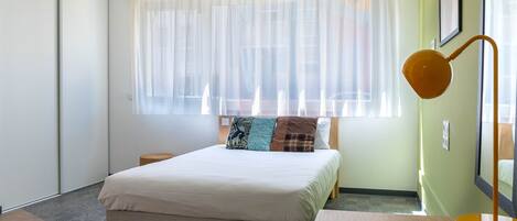 Un dormitorio con cama, mesita de noche, lámpara y ventana grande con cortinas ligeras.