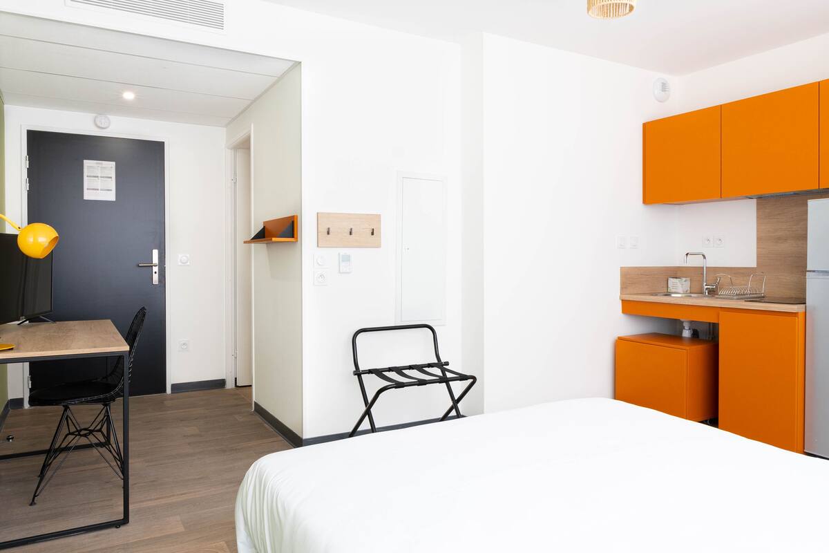 A compact room with a bed, a desk, a chair, and a kitchenette with orange cabinets.