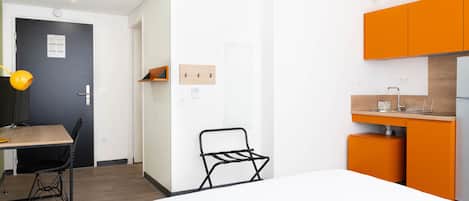 Ein gemütlicher Raum mit Bett, Schreibtisch, Stuhl und einer Küchenzeile mit orangenen Schränken.