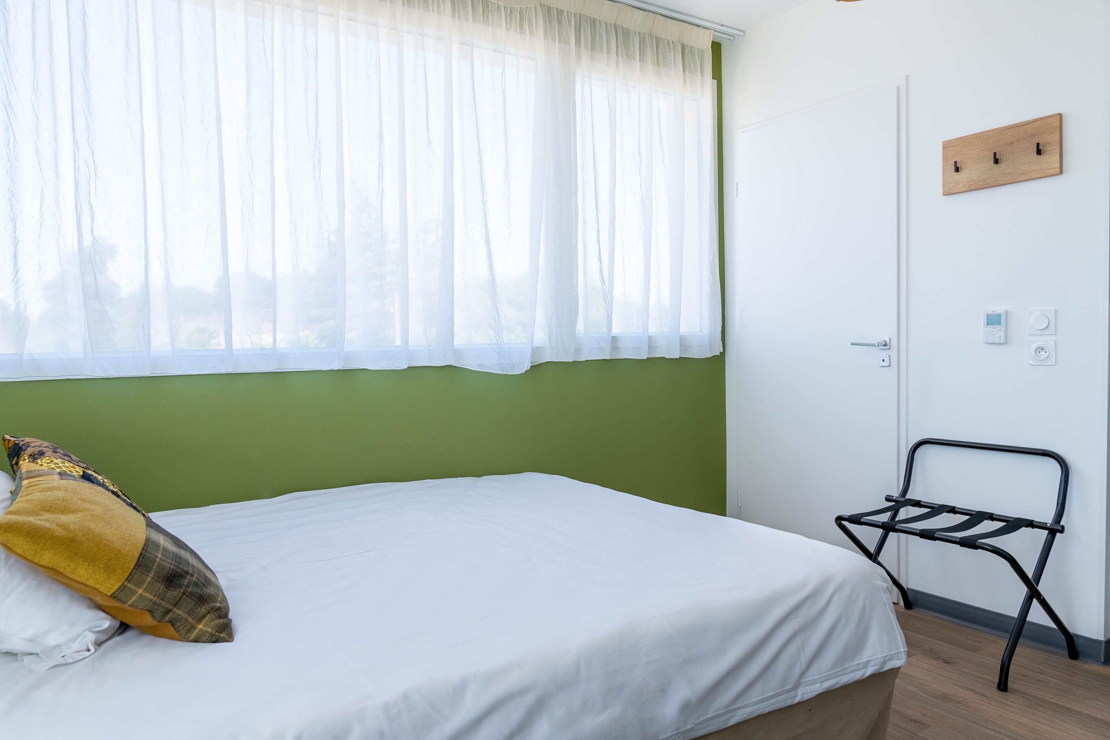 Quarto com cama, janela com cortinas leves, parede verde, cabideiro de madeira e porta com interruptor de luz.