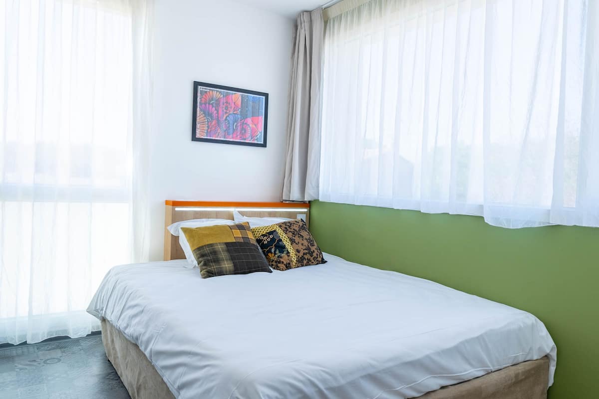 A neatly made bed with white bedding and multiple pillows, a framed artwork on the wall, and sheer curtains on the windows.