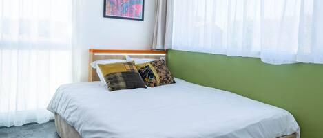 A neatly made bed with white bedding and multiple pillows, a framed artwork on the wall, and sheer curtains on the windows.