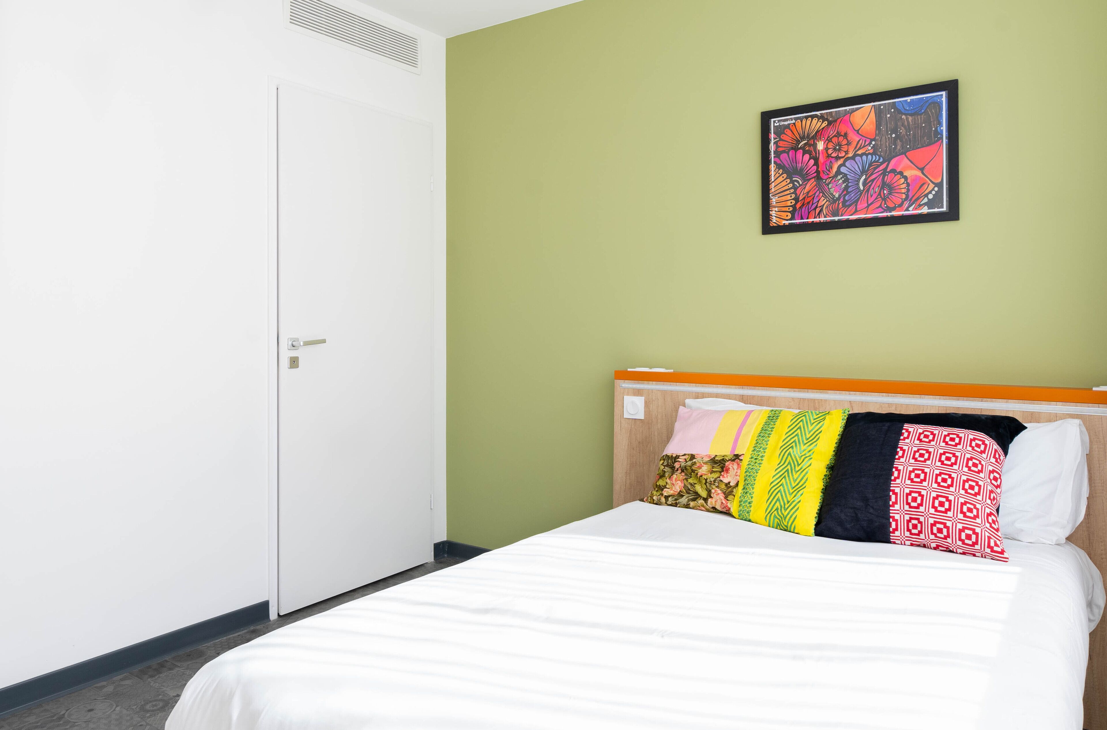A hotel room with a bed, a door, and a wall with a colorful painting.