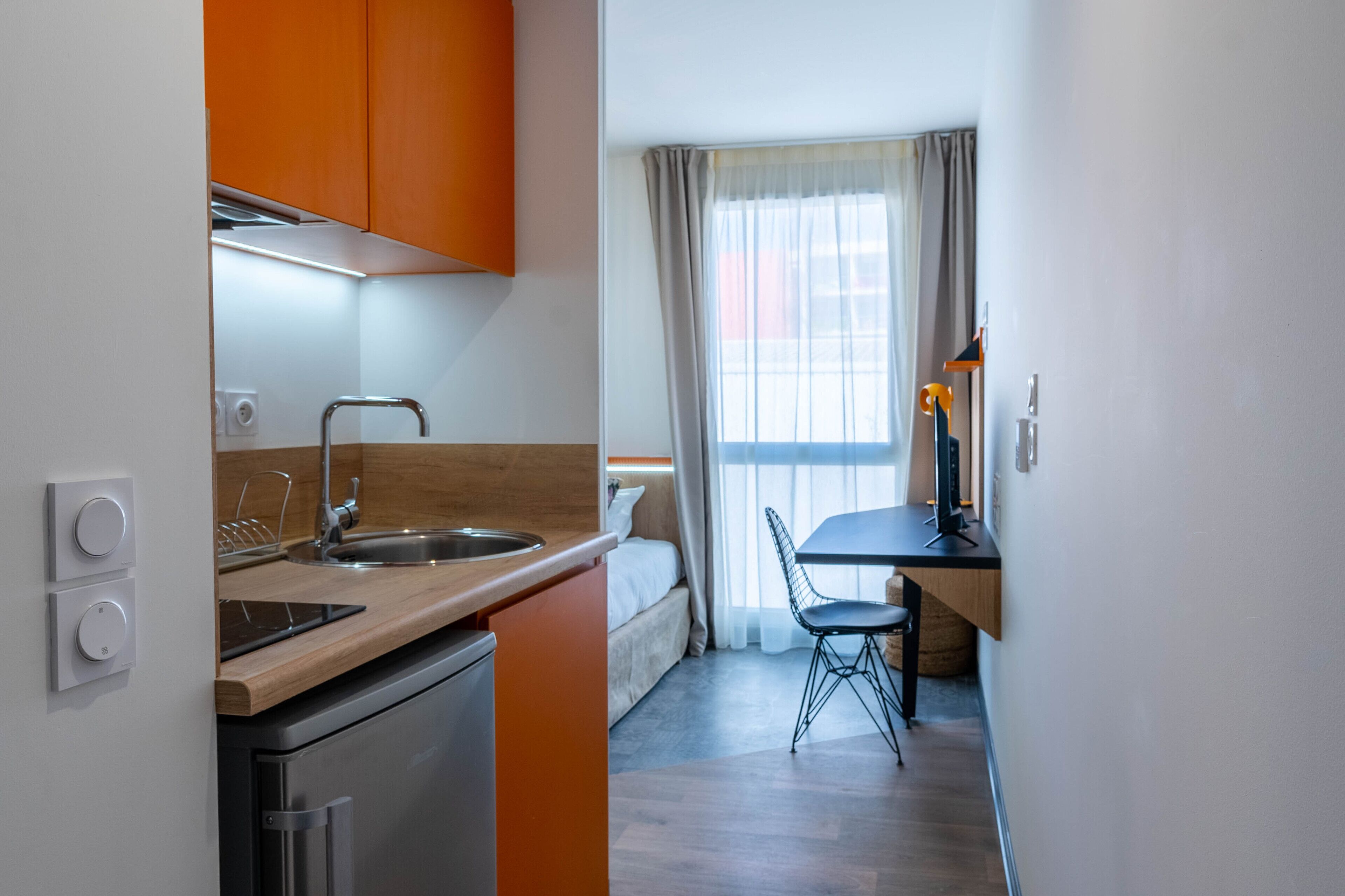 A compact kitchen with a sink, stove, and modern appliances, a dining area with a table and chair, and a bed in the background.