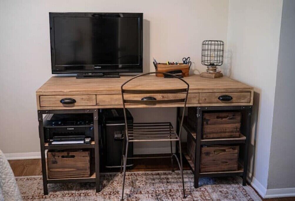 A wooden desk with a flat-screen TV, drawers, and a shelf unit with electronic devices.