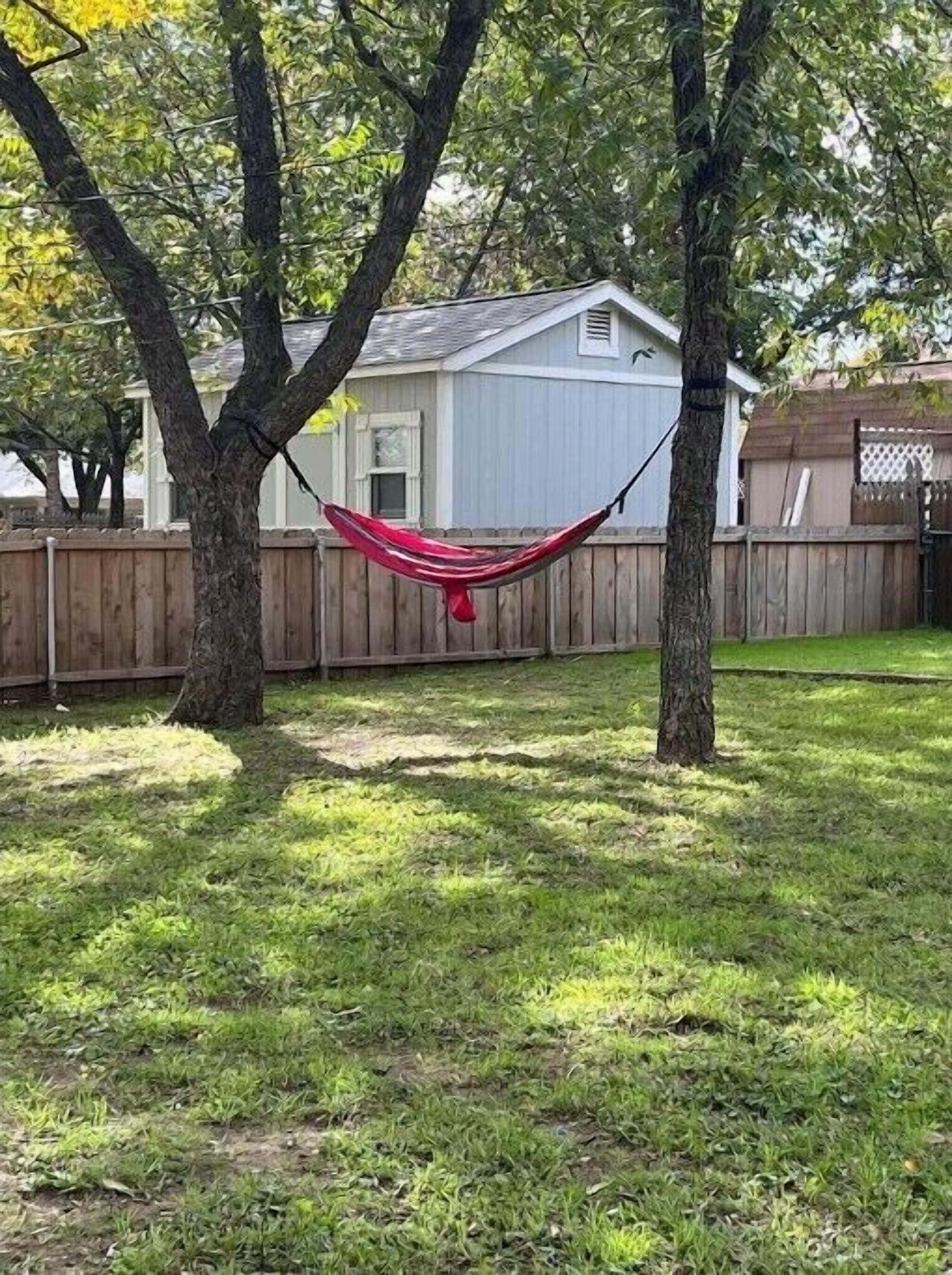 Una hamaca entre dos árboles en un patio trasero con una valla de madera y una casa al fondo.