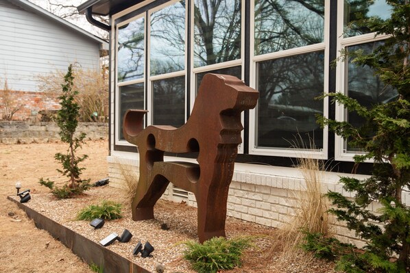 Un espace extérieur aménagé dans un style rustique, avec une sculpture en métal devant une maison dotée de grandes fenêtres.