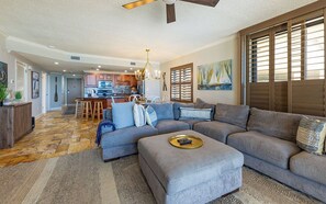 Ein modernes Wohnzimmer mit Ecksofa, Hocker und einem Essbereich, im Hintergrund ist eine Küche zu sehen.