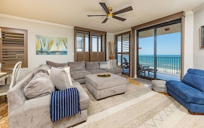 A living room with a sectional sofa, a blue armchair, a coffee table, a ceiling fan, and a view of the ocean.