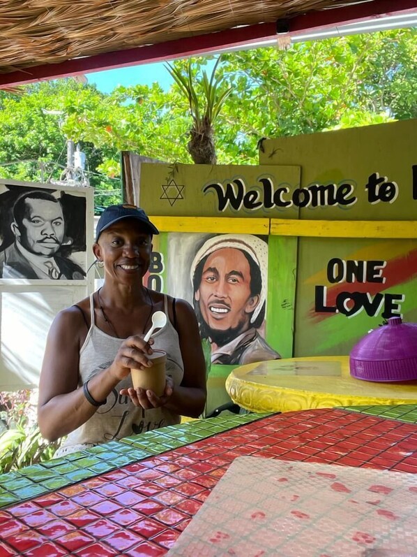 A person at a table with a colorful tiled floor, a 'Welcome to' sign, and a 'One Love' mural.