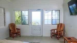 A room with a bed, two wooden chairs, a small table, a TV, and a view of the ocean through a door. - Oasis Palms Luxury Villa (Tower Isle)
