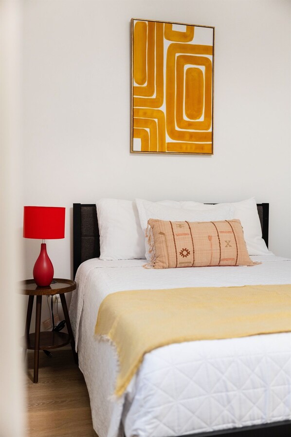 Un lit bien fait, avec du linge de lit blanc et un plaid jaune, une lampe rouge posée sur une table de chevet en bois, et une œuvre d’art aux motifs géométriques accrochée au mur.
