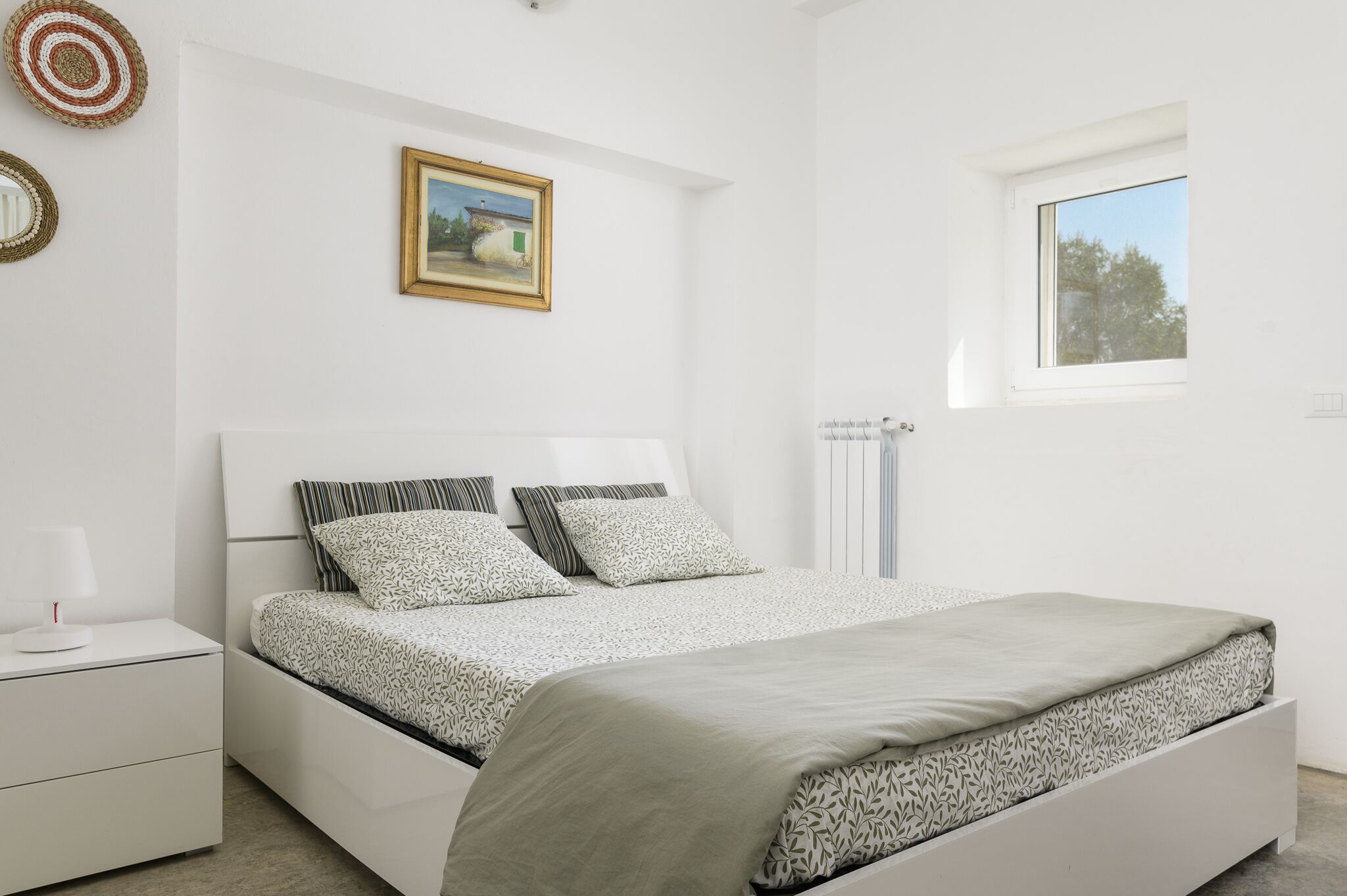 Ein Schlafzimmer mit einem Bett, einem Nachttisch, einem Heizkörper, einem Fenster und einem Wandbild.